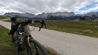 A bicycle is parked on a gravel path with a scenic background of a mountain range and cloudy skies. There are grassy fields on either side of the path, and a few houses are visible in the distance.