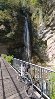 A serene waterfall cascading down a tall rock face surrounded by lush greenery. A bicycle is parked on a wooden bridge with metal railings, adding an element of exploration and adventure.