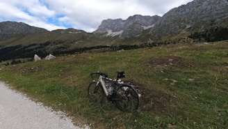 A bicycle is parked on a grassy, expansive plain with a backdrop of majestic mountains. The sky above is partly cloudy, softening the scene with a gentle light. The environment appears serene and inviting for outdoor activities like cycling or hiking.