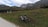 A bicycle is parked on a grassy, expansive plain with a backdrop of majestic mountains. The sky above is partly cloudy, softening the scene with a gentle light. The environment appears serene and inviting for outdoor activities like cycling or hiking.