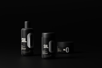 Three black cylindrical cosmetic containers with minimalist design and bold white lettering are set against a dark background. The containers vary in size and shape, including a tall bottle, a medium-sized jar, and a small jar. The lighting highlights the sleek, modern aesthetic of the products.