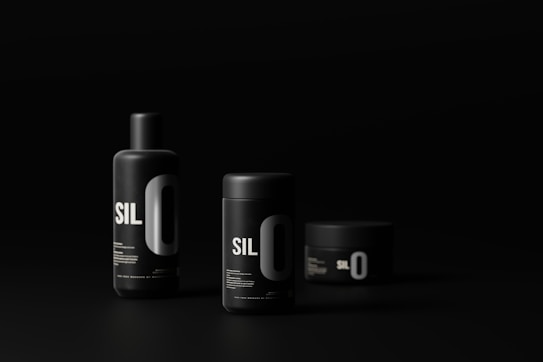 Three black cylindrical cosmetic containers with minimalist design and bold white lettering are set against a dark background. The containers vary in size and shape, including a tall bottle, a medium-sized jar, and a small jar. The lighting highlights the sleek, modern aesthetic of the products.