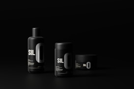 Three black cylindrical cosmetic containers with minimalist design and bold white lettering are set against a dark background. The containers vary in size and shape, including a tall bottle, a medium-sized jar, and a small jar. The lighting highlights the sleek, modern aesthetic of the products.