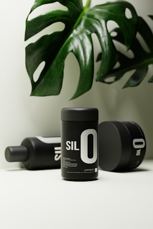 A minimalist setup featuring black cylindrical containers labeled 'SIL 0' with a glossy green monstera leaf overhead. The layout presents a clean, sophisticated aesthetic with a focus on the product design and natural element.