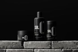 Three sleek black containers with minimalistic design are arranged on a stone-like surface. Each container has the bold text 'SIL O' printed on it, suggesting a cohesive branding. The background is a solid dark color, enhancing the modern and sophisticated look of the products.