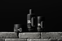 Three sleek black containers with minimalistic design are arranged on a stone-like surface. Each container has the bold text 'SIL O' printed on it, suggesting a cohesive branding. The background is a solid dark color, enhancing the modern and sophisticated look of the products.