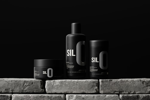 Three sleek black containers with minimalistic design are arranged on a stone-like surface. Each container has the bold text 'SIL O' printed on it, suggesting a cohesive branding. The background is a solid dark color, enhancing the modern and sophisticated look of the products.
