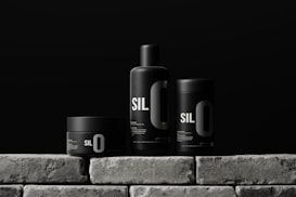Three sleek black containers with minimalistic design are arranged on a stone-like surface. Each container has the bold text 'SIL O' printed on it, suggesting a cohesive branding. The background is a solid dark color, enhancing the modern and sophisticated look of the products.