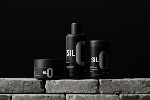 Three sleek black containers with minimalistic design are arranged on a stone-like surface. Each container has the bold text 'SIL O' printed on it, suggesting a cohesive branding. The background is a solid dark color, enhancing the modern and sophisticated look of the products.