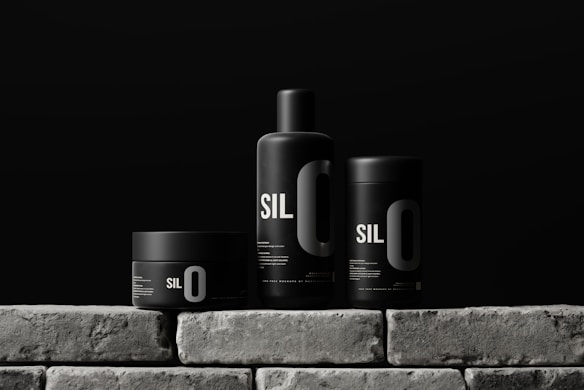 Three sleek black containers with minimalistic design are arranged on a stone-like surface. Each container has the bold text 'SIL O' printed on it, suggesting a cohesive branding. The background is a solid dark color, enhancing the modern and sophisticated look of the products.