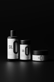 Three sleek, minimalist containers with black lids and bold white labels, arranged against a dark background. The labels feature large, prominent text reading 'SIL O' with smaller text beneath.