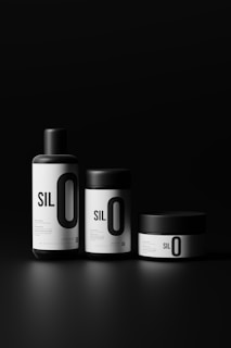 Three sleek, minimalist containers with black lids and bold white labels, arranged against a dark background. The labels feature large, prominent text reading 'SIL O' with smaller text beneath.