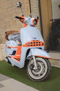 A stylish scooter with a modern design features a light blue body with vibrant orange accents. The front showcases a sleek headlight and handlebars, paired with shiny chrome mirrors. The scooter is parked on a patch of artificial grass against a textured beige brick wall. A brown leather bag is attached to the back of the seat.