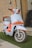 A stylish scooter with a modern design features a light blue body with vibrant orange accents. The front showcases a sleek headlight and handlebars, paired with shiny chrome mirrors. The scooter is parked on a patch of artificial grass against a textured beige brick wall. A brown leather bag is attached to the back of the seat.