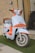 A stylish scooter with a modern design features a light blue body with vibrant orange accents. The front showcases a sleek headlight and handlebars, paired with shiny chrome mirrors. The scooter is parked on a patch of artificial grass against a textured beige brick wall. A brown leather bag is attached to the back of the seat.