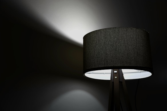 A modern, black lampshade on a tripod stand is illuminated against a dark background. The light casts a soft glow and distinct shadow patterns on the wall behind it, creating a dramatic contrast.