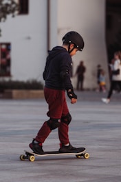 A child wearing protective gear, including a helmet and elbow and knee pads, is riding a skateboard in an urban setting. The child is dressed in a dark hoodie and maroon pants, focusing on balancing the skateboard. The background shows a few blurred figures, suggesting a public area.