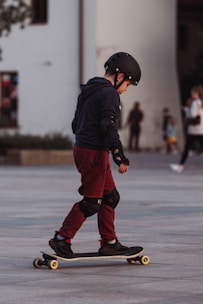 A child wearing protective gear, including a helmet and elbow and knee pads, is riding a skateboard in an urban setting. The child is dressed in a dark hoodie and maroon pants, focusing on balancing the skateboard. The background shows a few blurred figures, suggesting a public area.