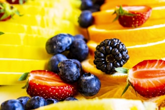 A vibrant assortment of fresh fruits, including sliced strawberries, blueberries, blackberries, and orange slices, arranged closely together. The image captures the juicy textures and vivid colors of the fruits, suggesting freshness and vitality.