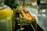 An elegantly arranged display of gourmet appetizers on a glass-topped table. The assortment includes bite-sized treats featuring colorful garnishes such as red caviar, blueberries, and herbs. The table has a gold metallic frame and blurred background, indicating a setting with sophisticated decor.