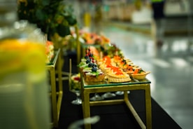 An elegantly arranged display of gourmet appetizers on a glass-topped table. The assortment includes bite-sized treats featuring colorful garnishes such as red caviar, blueberries, and herbs. The table has a gold metallic frame and blurred background, indicating a setting with sophisticated decor.