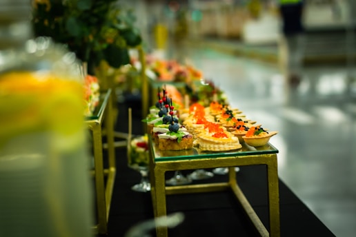 An elegantly arranged display of gourmet appetizers on a glass-topped table. The assortment includes bite-sized treats featuring colorful garnishes such as red caviar, blueberries, and herbs. The table has a gold metallic frame and blurred background, indicating a setting with sophisticated decor.