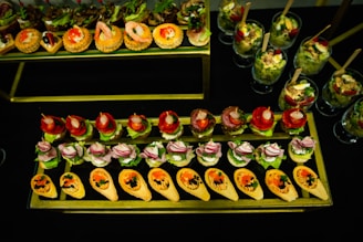 An assortment of elegantly arranged canapés is displayed on a tiered serving tray, featuring a variety of toppings like shrimp, radishes, and greens. The setup includes a mix of savory tartlets and stuffed shells, with a colorful selection of garnishes. Several glasses with layered salads and wooden forks are placed nearby, contributing to the diverse presentation.