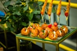 A display of appetizers arranged on a glass and gold-framed table. There are mini croissants filled with different ingredients such as cream cheese and vegetables, garnished with small skewers holding items like olives and tomatoes. Behind them, shrimp cocktails are served in tall glasses. This takes place against a backdrop of green plants.