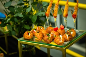 A display of appetizers arranged on a glass and gold-framed table. There are mini croissants filled with different ingredients such as cream cheese and vegetables, garnished with small skewers holding items like olives and tomatoes. Behind them, shrimp cocktails are served in tall glasses. This takes place against a backdrop of green plants.