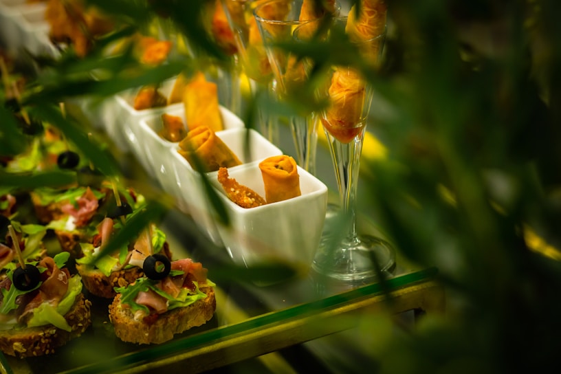An arrangement of small gourmet appetizers placed on a buffet table includes toasted bread slices topped with lettuce, sliced ham, and black olives, as well as crispy wrapped spring rolls displayed in elegant white trays and wine glasses. The vibrant green leaves partially obscure the view, providing a lush framing for the scene.