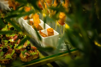An arrangement of small gourmet appetizers placed on a buffet table includes toasted bread slices topped with lettuce, sliced ham, and black olives, as well as crispy wrapped spring rolls displayed in elegant white trays and wine glasses. The vibrant green leaves partially obscure the view, providing a lush framing for the scene.