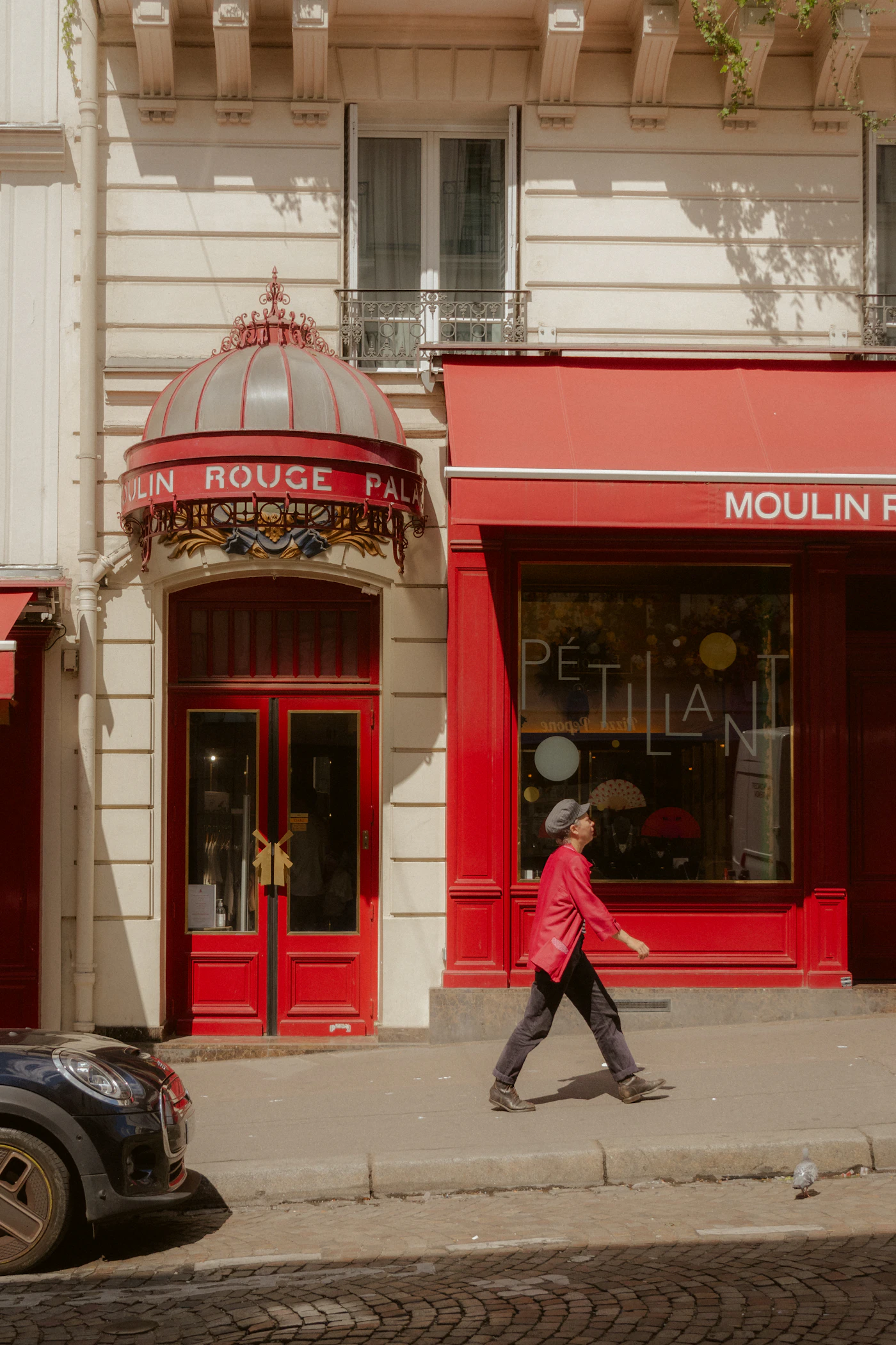 The red painted frontage of the Moulin Rouge by day, a person walking past on the pavement
