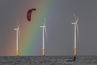 Three large wind turbines stand in a body of water with a vibrant rainbow in the background. A person is kite surfing near the turbines, with a red kite visible above.