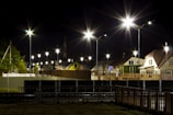 A nighttime scene featuring a well-lit residential street with modern houses and bright streetlights. The streetlights create starburst effects against the dark sky. The area is fenced with metal barriers, and there is a small patch of grass in the foreground.