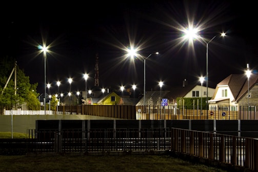 A nighttime scene featuring a well-lit residential street with modern houses and bright streetlights. The streetlights create starburst effects against the dark sky. The area is fenced with metal barriers, and there is a small patch of grass in the foreground.