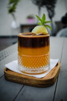 A beautifully crafted drink in a textured glass, resting on a wooden coaster on a grey table. The drink has a dark and amber layer topped with a slice of lime and a green garnish.