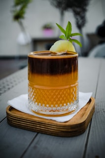 A beautifully crafted drink in a textured glass, resting on a wooden coaster on a grey table. The drink has a dark and amber layer topped with a slice of lime and a green garnish.