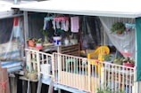 A small balcony area decorated with various potted plants, hanging clothes, and household items. The space features a yellow plastic chair, a wooden crate with containers, and colorful textiles. The setting is modest and informal, suggesting a personal and lived-in vibe.