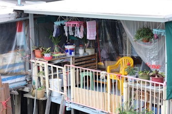 A small balcony area decorated with various potted plants, hanging clothes, and household items. The space features a yellow plastic chair, a wooden crate with containers, and colorful textiles. The setting is modest and informal, suggesting a personal and lived-in vibe.