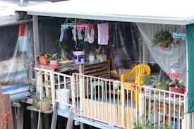 A small balcony area decorated with various potted plants, hanging clothes, and household items. The space features a yellow plastic chair, a wooden crate with containers, and colorful textiles. The setting is modest and informal, suggesting a personal and lived-in vibe.