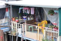 A small balcony area decorated with various potted plants, hanging clothes, and household items. The space features a yellow plastic chair, a wooden crate with containers, and colorful textiles. The setting is modest and informal, suggesting a personal and lived-in vibe.