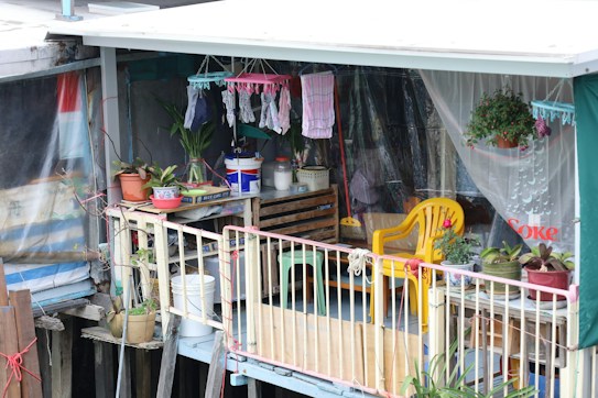 A small balcony area decorated with various potted plants, hanging clothes, and household items. The space features a yellow plastic chair, a wooden crate with containers, and colorful textiles. The setting is modest and informal, suggesting a personal and lived-in vibe.