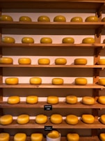 A collection of large, round wheels of cheese neatly arranged on wooden shelves. The cheeses are uniformly golden-yellow, and each shelf holds a row of them. Small signs with prices are placed near some of the cheeses.