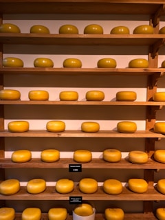 A collection of large, round wheels of cheese neatly arranged on wooden shelves. The cheeses are uniformly golden-yellow, and each shelf holds a row of them. Small signs with prices are placed near some of the cheeses.