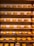 A collection of large, round wheels of cheese neatly arranged on wooden shelves. The cheeses are uniformly golden-yellow, and each shelf holds a row of them. Small signs with prices are placed near some of the cheeses.