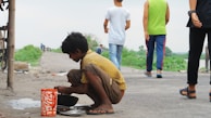 A young boy squats on a dirt path, cleaning kitchen utensils next to a bright orange and white patterned container. The boy is focused on his task, wearing a yellow spotted shirt and brown shorts. In the background, several people walk along the path, with greenery and a cloudy sky visible.
