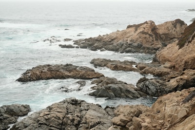 A rugged coastline with waves crashing against weathered rocks under a cloudy sky.