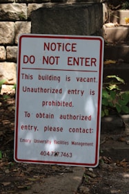 A warning sign is placed outdoors against a background of stone steps and walls, surrounded by some greenery. The sign indicates that the building is vacant and unauthorized entry is prohibited. Contact information for further inquiries is provided at the bottom.