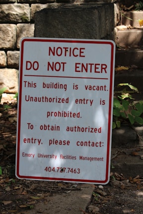 A warning sign is placed outdoors against a background of stone steps and walls, surrounded by some greenery. The sign indicates that the building is vacant and unauthorized entry is prohibited. Contact information for further inquiries is provided at the bottom.