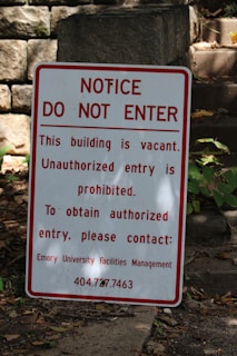 A warning sign is placed outdoors against a background of stone steps and walls, surrounded by some greenery. The sign indicates that the building is vacant and unauthorized entry is prohibited. Contact information for further inquiries is provided at the bottom.